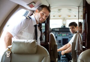 Two pilots in a private jet's cockpit, one smiling and standing by the doorway while the other is seated at the controls, showcasing a team ready for flight.