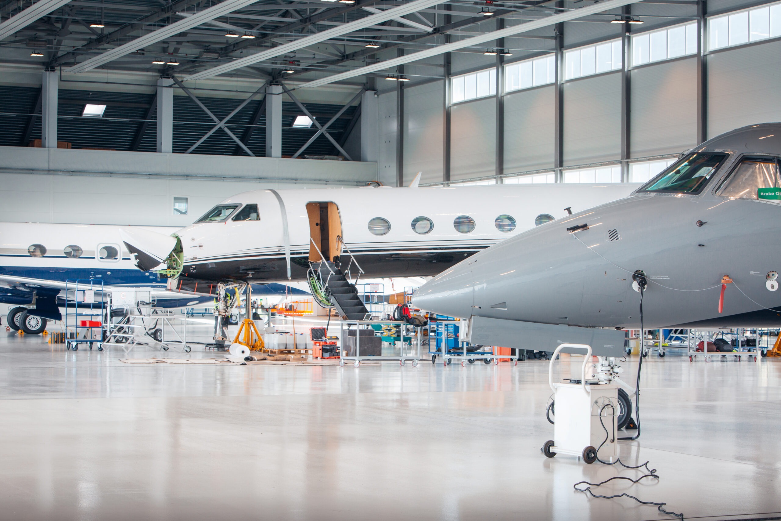 Two aircraft are inside a well-lit hangar, with one airplane undergoing maintenance. Tools and equipment are visible, highlighting an active aviation service environment.